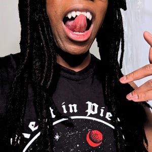 May include: A person with dreadlocks wearing a black t-shirt with the text "t in Pie" and a celestial design. The person is showing their open mouth with vampire fangs.