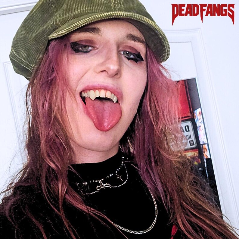 May include: A person with pink hair and dark eye makeup, wearing a green corduroy cap and a black top. The person is sticking out their tongue and showing vampire fangs. A silver necklace is around their neck. The text "DEADFANGS" is in the upper right corner.
