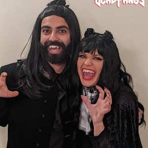 May include: Two people dressed in vampire costumes, with black wigs, capes, and fake fangs. The woman wears a white ruffled shirt and a black choker. The man has a beard and a black coat. The text "DEADFANGS" is at the top.