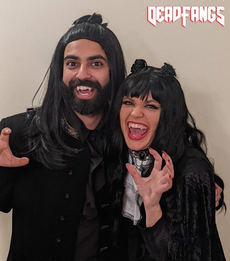 May include: Two people dressed in vampire costumes, with black wigs, capes, and fake fangs. The woman wears a white ruffled shirt and a black choker. The man has a beard and a black coat. The text "DEADFANGS" is at the top.