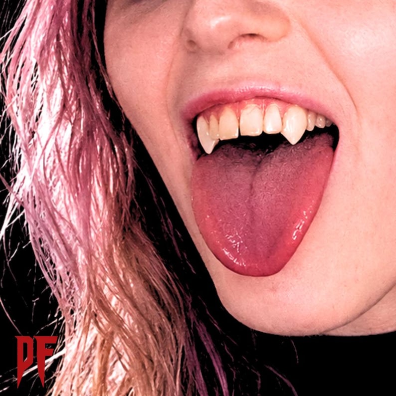May include: Close-up of a mouth with sharp, pointed vampire fangs. The tongue is extended, and the lips are parted. The person has pink hair. The image has a dark background.