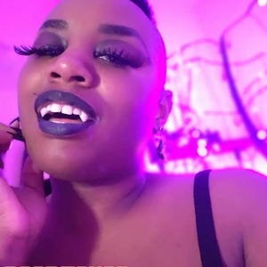 May include: A person with dark makeup, including dark lipstick and long eyelashes, wearing vampire fangs. The text "DEADFANGS" is displayed in a bold, stylized font at the bottom of the image.