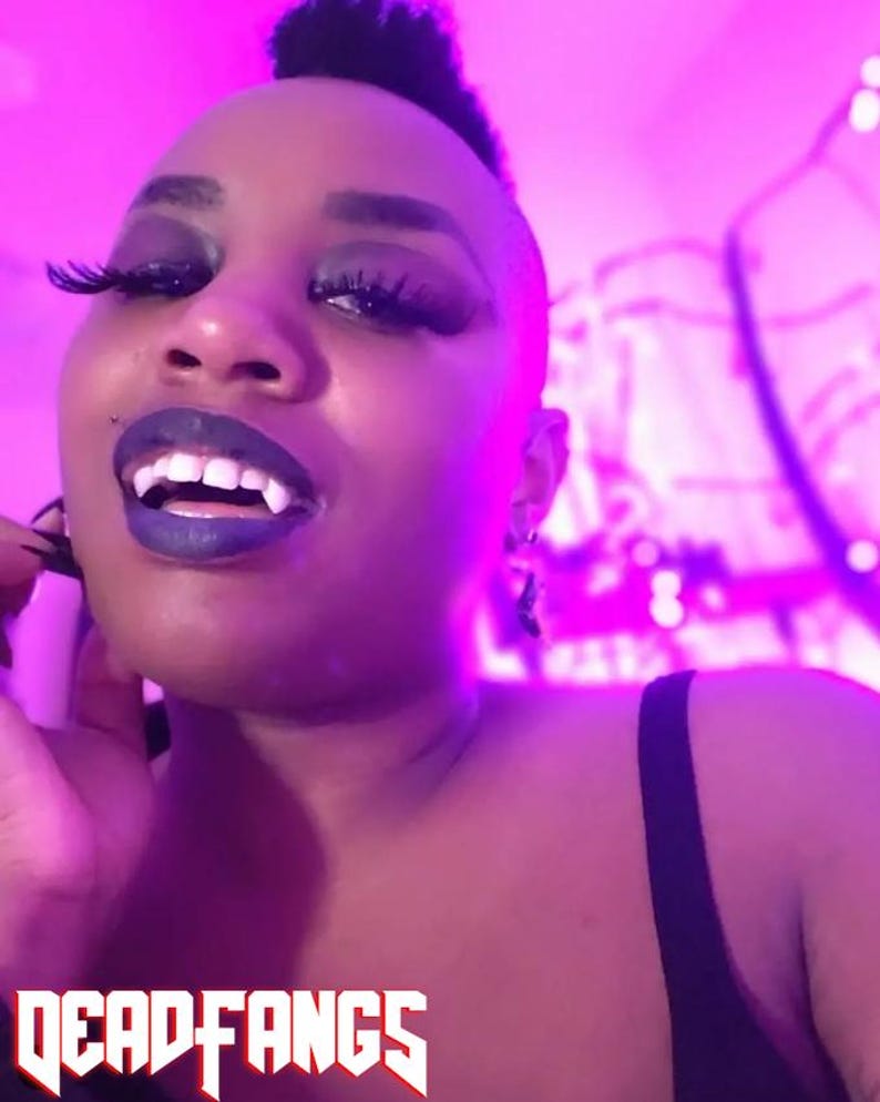 May include: A person with dark makeup, including dark lipstick and long eyelashes, wearing vampire fangs. The text "DEADFANGS" is displayed in a bold, stylized font at the bottom of the image.