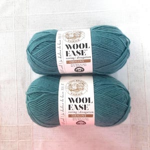 Peut inclure: Deux pelotes de laine bleu-vert Lion Brand Wool-Ease Origins. Chaque pelote porte une étiquette blanche avec le nom de la marque, le type de fil et les détails d'origine. La laine est de style roving, idéale pour le tricot ou le crochet.