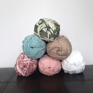 May include: A pyramid of seven yarn balls in various colors. The colors include green, blue, tan, pink, and white. The yarn is thick and appears to be cotton or a similar fiber. The balls are stacked on a dark surface against a white background.