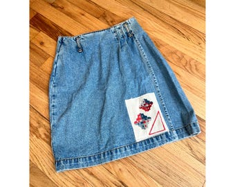 May include: Light blue denim skirt with a decorative patch. The patch features red, white, and blue cross-stitch designs and a red triangle. The skirt has a classic five-pocket design and a button and zipper closure.