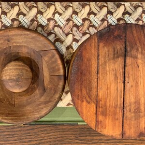 Op de afbeelding: Twee ronde houten deksels, een met een gat in het midden en de andere met een glad oppervlak. Beide deksels zijn donkerbruin van kleur en hebben een rustieke uitstraling.