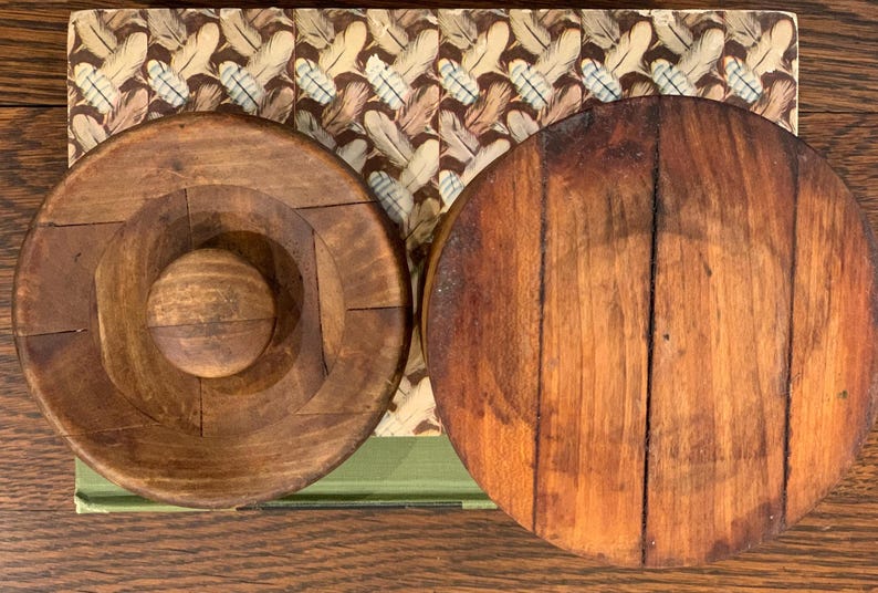 Op de afbeelding: Twee ronde houten deksels, een met een gat in het midden en de andere met een glad oppervlak. Beide deksels zijn donkerbruin van kleur en hebben een rustieke uitstraling.