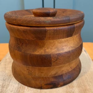 Op de afbeelding: Een houten container met een deksel. De container is gemaakt van licht en donkerbruin hout en heeft een afgeronde vorm. Het deksel heeft een kleine knop bovenop.