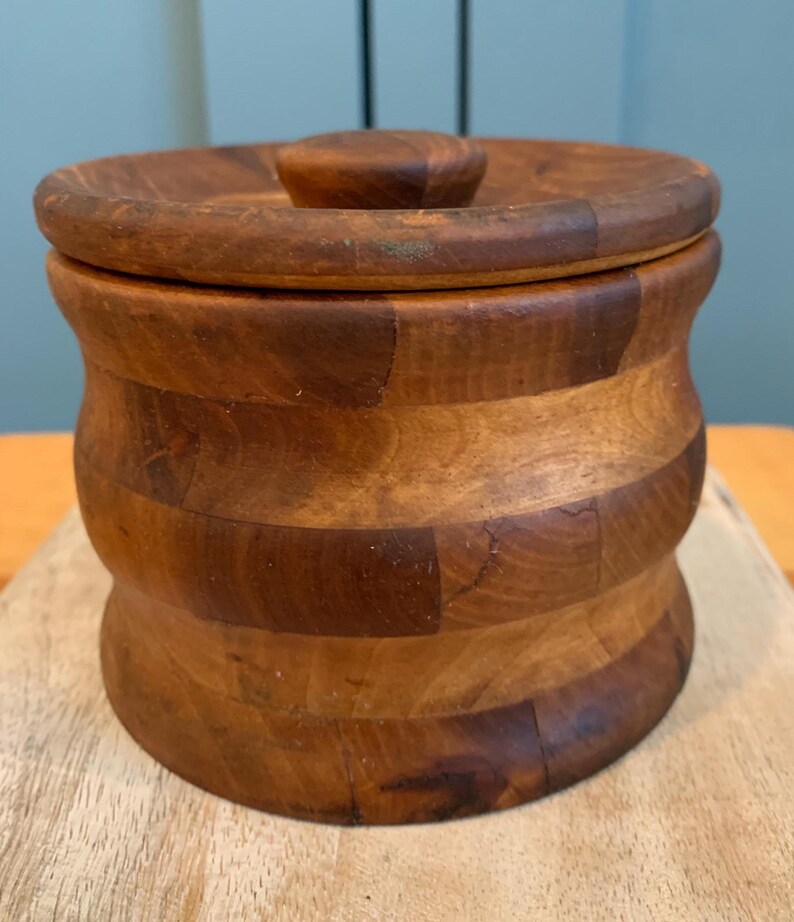 Op de afbeelding: Een houten container met een deksel. De container is gemaakt van licht en donkerbruin hout en heeft een afgeronde vorm. Het deksel heeft een kleine knop bovenop.