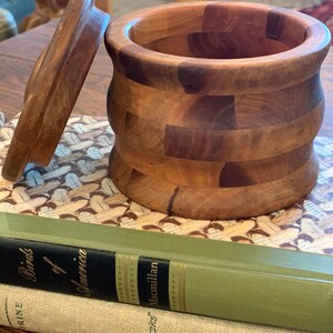 Op de afbeelding: Een houten container met een deksel, gemaakt van meerdere stukken hout, met een natuurlijke houtkleur. De container staat op een boek met de titel "Birds of America" op de omslag.
