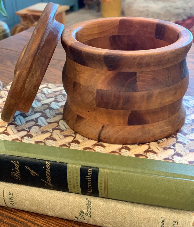 Op de afbeelding: Een houten container met een deksel, gemaakt van meerdere stukken hout, met een natuurlijke houtkleur. De container staat op een boek met de titel "Birds of America" op de omslag.