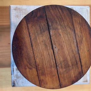 Op de afbeelding: Een ronde houten tafelblad met een donkerbruine vlek. De houtnerf is zichtbaar en er zitten verschillende knopen in het hout.