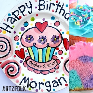 May include: A white ceramic plate with a hand-painted cupcake design. The cupcake is pink with green and blue frosting and has three purple flowers on top. The plate also has the words "Happy Birthday" and "Morgan" written on it, along with the date "October 9, 2019".