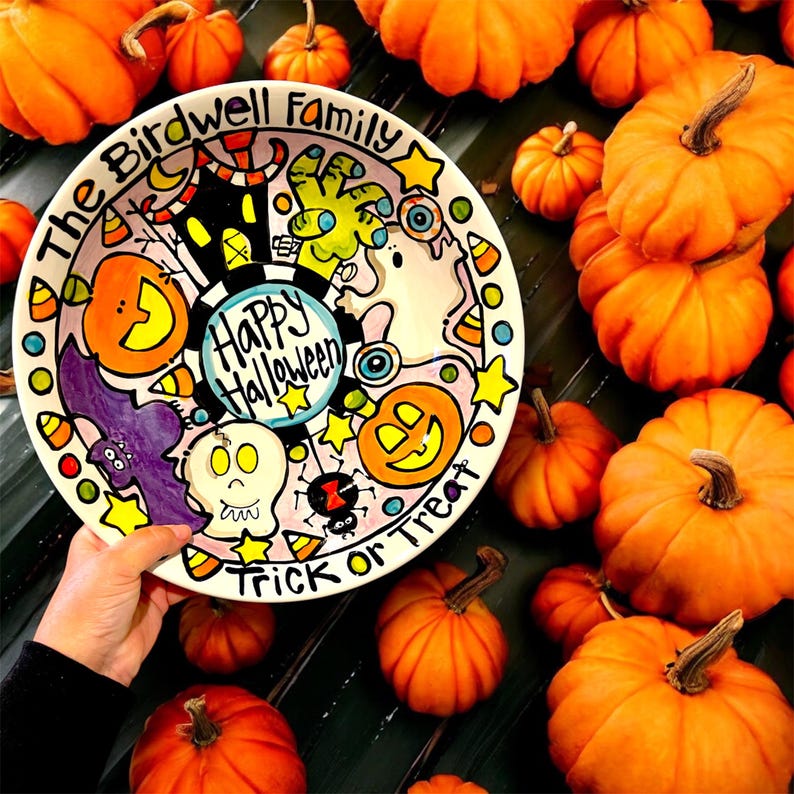 May include: A round ceramic plate featuring a hand-painted Halloween theme with a central Happy Halloween text, surrounded by illustrations of a pumpkin, a skull, a bat, a spider, a ghost, and a zombie hand, with The Birdwell Family Trick or Treat text.
