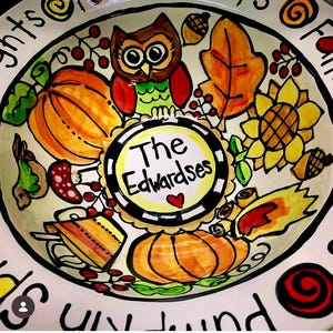 May include: A round ceramic bowl featuring a hand-painted autumn theme with pumpkins, an owl, a sunflower, leaves, and acorns. The center circle contains the text The Edwardsets with a small red heart. The rim displays partial black text.