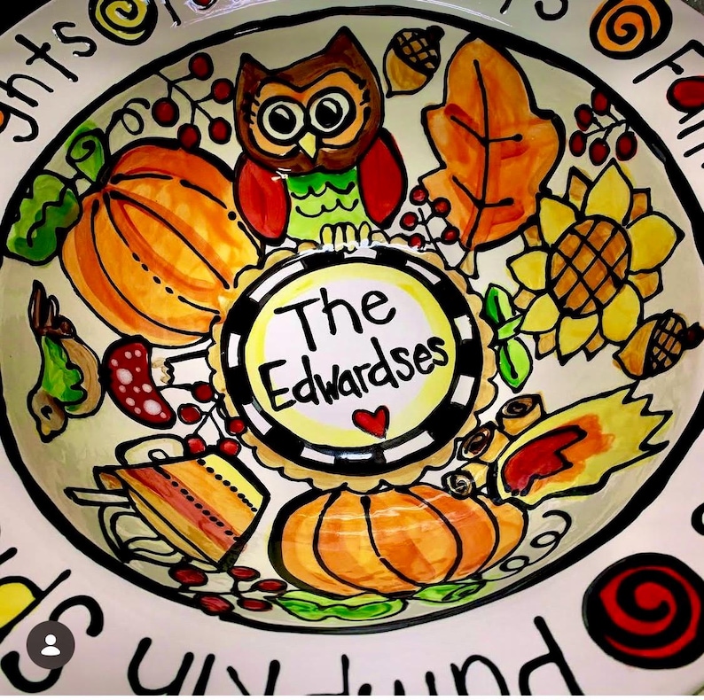 May include: A round ceramic bowl featuring a hand-painted autumn theme with pumpkins, an owl, a sunflower, leaves, and acorns. The center circle contains the text The Edwardsets with a small red heart. The rim displays partial black text.
