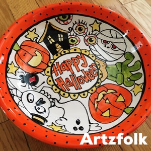 May include: A round ceramic bowl with an orange rim featuring black dots. The center displays the text Happy Halloween surrounded by illustrations of a black house, eyeballs, a mummy, a green hand, pumpkins, a spider, a skull, and a ghost.