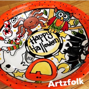 May include: A round ceramic plate with an orange rim featuring black dots. The center displays the text Happy Halloween surrounded by hand-painted illustrations of a brown spider, a colorful owl, a black bat, a white ghost, and a pumpkin.