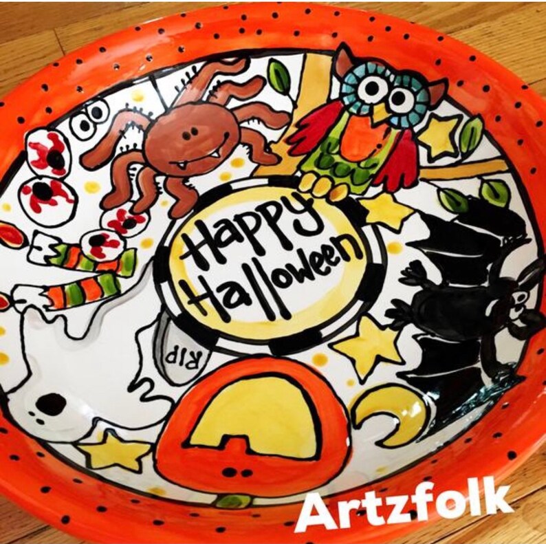 May include: A round ceramic plate with an orange rim featuring black dots. The center displays the text Happy Halloween surrounded by hand-painted illustrations of a brown spider, a colorful owl, a black bat, a white ghost, and a pumpkin.