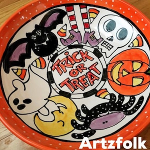 May include: A round ceramic bowl with an orange rim featuring white polka dots. The center displays the text "TRICK OR TREAT" surrounded by hand-painted Halloween illustrations including a black bat, a skull, a pumpkin, a ghost, a spider, and candy corn.