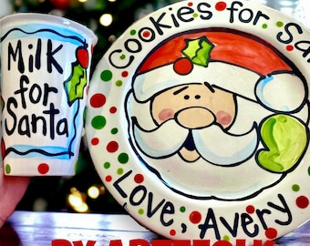 May include: A hand-painted ceramic plate and cup set for Santa. The plate features a Santa Claus face with the words "Cookies for Santa" and "Love, Avery". The cup says "Milk for Santa". The set is decorated with red, green, and white accents.