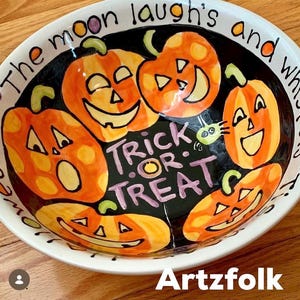 May include: A round ceramic bowl with a black interior featuring five orange jack-o'-lanterns and the purple text "TRICK OR TREAT". The white rim displays the text "The moon laugh's and whispers". The bowl has a glossy finish and hand-painted details.