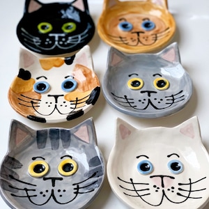 May include: Six small ceramic cat dishes in various colors and patterns. Each dish features a cat face with large eyes and a smiling mouth. The dishes are perfect for serving small snacks or treats to cats.