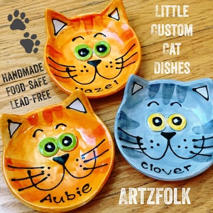 May include: Three handmade ceramic cat dishes with whimsical cat faces. The dishes are orange, blue, and yellow with black outlines and large googly eyes. The dishes are labeled "Aubie", "Mazel", and "Clover".