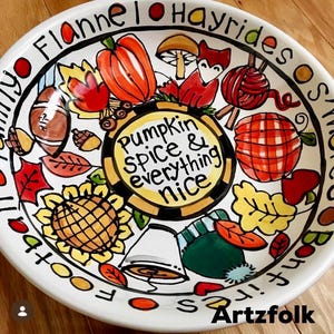 May include: A white ceramic bowl featuring hand-painted autumn illustrations including a pumpkin, fox, sunflower, football, and leaves. The center text reads, "Pumpkin spice & everything nice," while the rim text includes "Flannel," "Hayrides," and "Bonfires."