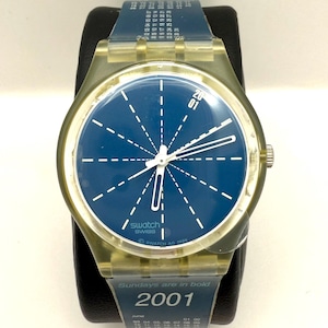 May include: A Swatch watch with a transparent case and a blue dial featuring white lines radiating from the center. The watch band is blue and displays the year 2001 and a calendar. The watch is displayed on a black stand.