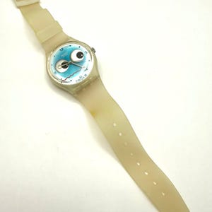 May include: A vintage Swatch watch with a light blue face featuring a cartoon eye design. The watch has a cream-colored, transparent plastic strap and a silver-tone case. The watch band is approximately 9 inches long.