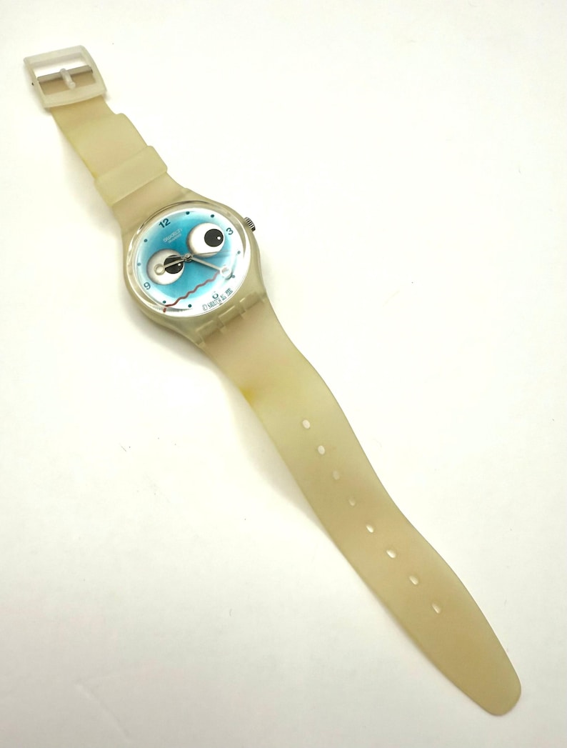 May include: A vintage Swatch watch with a light blue face featuring a cartoon eye design. The watch has a cream-colored, transparent plastic strap and a silver-tone case. The watch band is approximately 9 inches long.