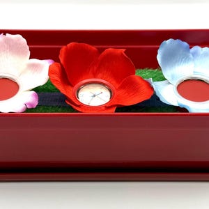 May include: A burgundy rectangular planter with three artificial flowers: white, red, and blue. Each flower has a red centre. The planter sits on a matching tray. The planter is approximately 30 cm long.