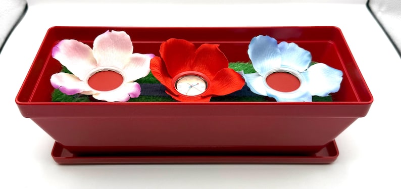 May include: A burgundy rectangular planter with three artificial flowers: white, red, and blue. Each flower has a red centre. The planter sits on a matching tray. The planter is approximately 30 cm long.