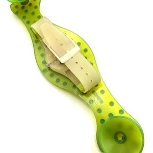 May include: A transparent watch with a cream-colored band and a green, polka-dotted holder. The watch is secured by a cream-colored buckle. The holder has two green suction cups on each end.