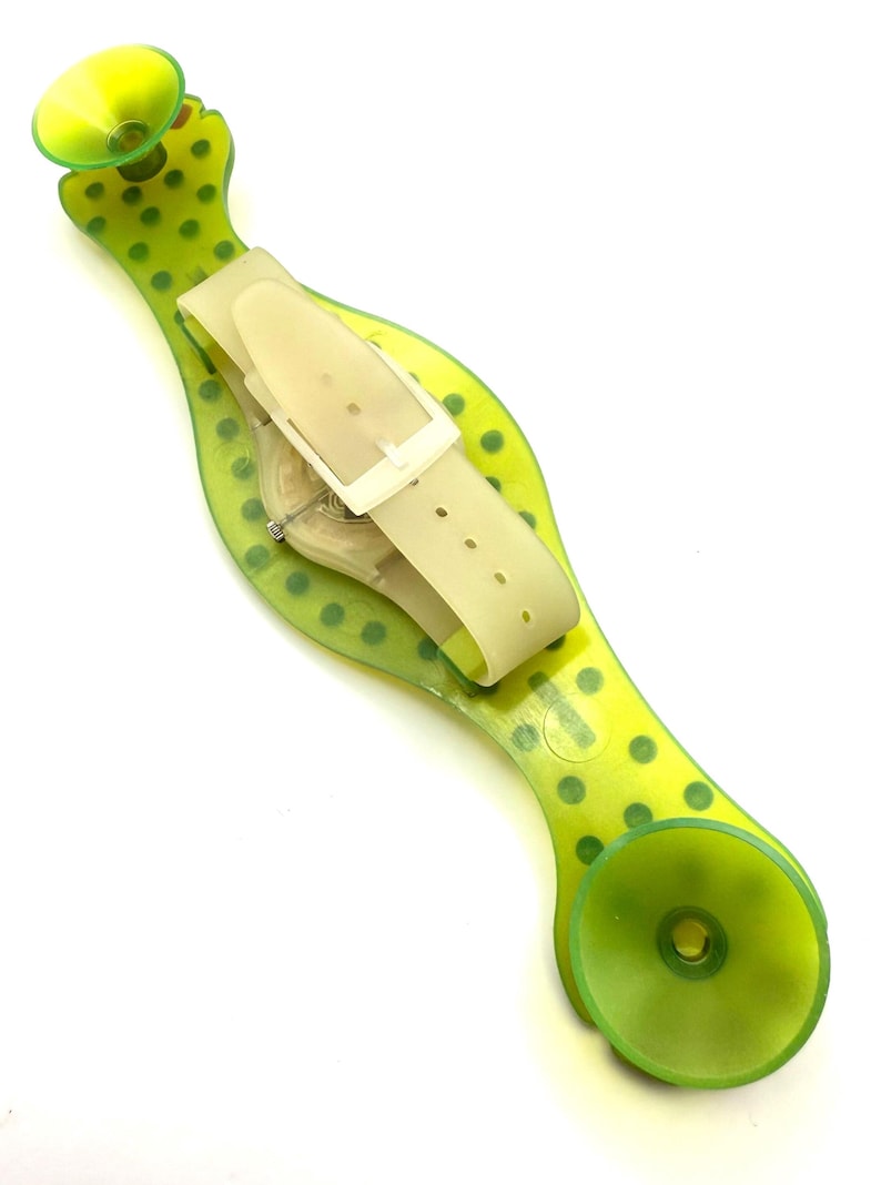 May include: A transparent watch with a cream-colored band and a green, polka-dotted holder. The watch is secured by a cream-colored buckle. The holder has two green suction cups on each end.