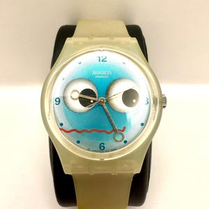 May include: A Swatch watch with a whimsical design. The watch face features a blue background with large cartoon eyes, a red mouth, and the text "Swatch Swiss". The watch has a clear, light-colored plastic case and band.
