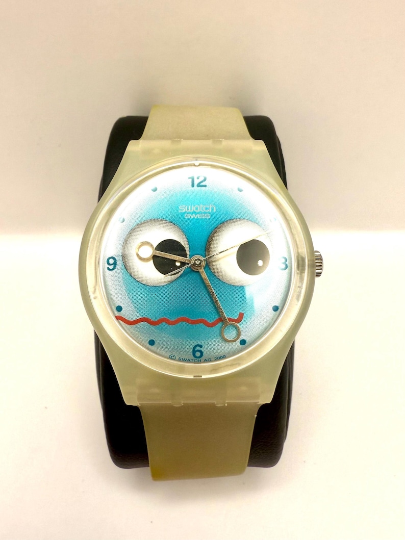 May include: A Swatch watch with a whimsical design. The watch face features a blue background with large cartoon eyes, a red mouth, and the text "Swatch Swiss". The watch has a clear, light-colored plastic case and band.