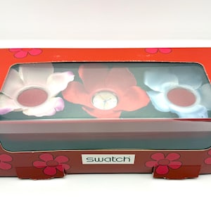 May include: A red box with a clear window showing three flower-shaped watches. The box is decorated with a floral pattern and the brand name "Swatch". The watches have pink, red, and blue petals.