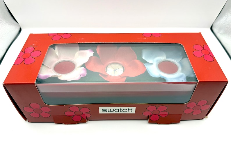 May include: A red box with a clear window showing three flower-shaped watches. The box is decorated with a floral pattern and the brand name "Swatch". The watches have pink, red, and blue petals.
