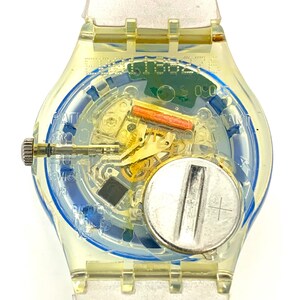 Pu&ograve; includere: Un orologio Swatch trasparente con cassa e cinturino giallo trasparente. Il quadrante rivela il meccanismo interno, tra cui una batteria, ingranaggi e un quadrante con motivo blu e bianco. L'orologio ha una corona argentata e un cinturino trasparente.