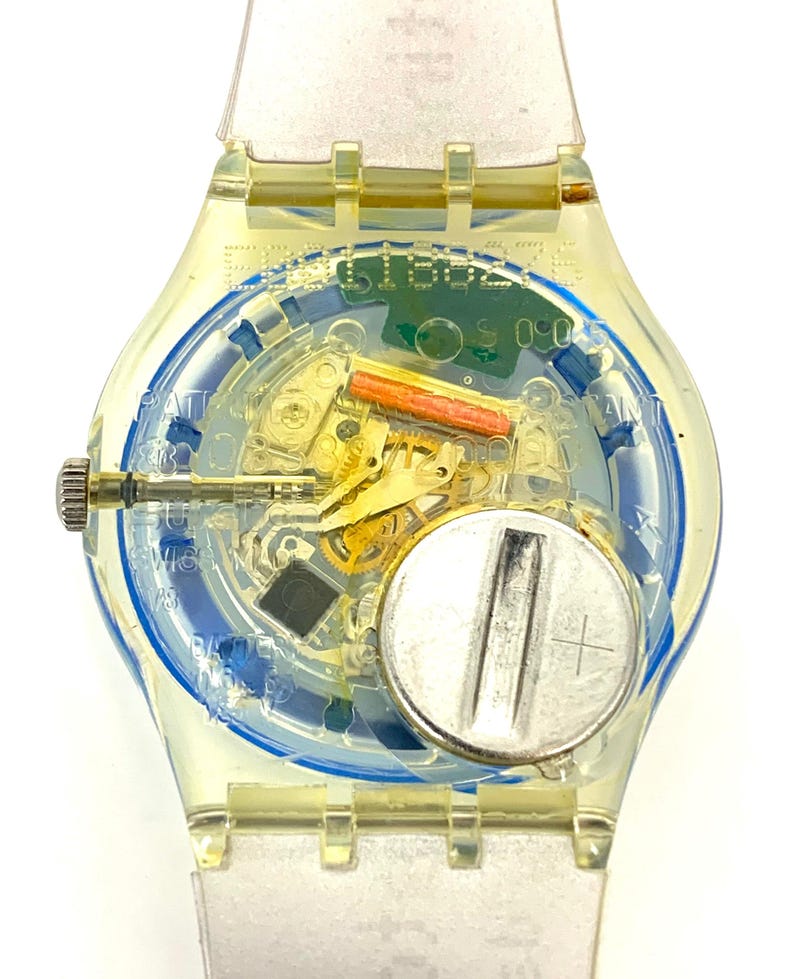 Pu&ograve; includere: Un orologio Swatch trasparente con cassa e cinturino giallo trasparente. Il quadrante rivela il meccanismo interno, tra cui una batteria, ingranaggi e un quadrante con motivo blu e bianco. L'orologio ha una corona argentata e un cinturino trasparente.