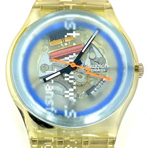 Pu&ograve; includere: Un orologio Swatch Swiss trasparente con un anello del quadrante blu chiaro e lancette arancioni. Il quadrante dell'orologio mostra il meccanismo interno. Il cinturino dell'orologio riporta le parole "setting" e "date".