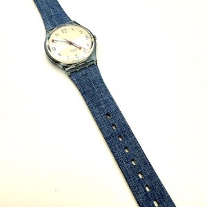 May include: A wristwatch with a clear plastic case and a denim-style blue strap. The watch face is off-white with black numerals and red and blue hands. The strap has a buckle closure and measures approximately 23 cm in length.