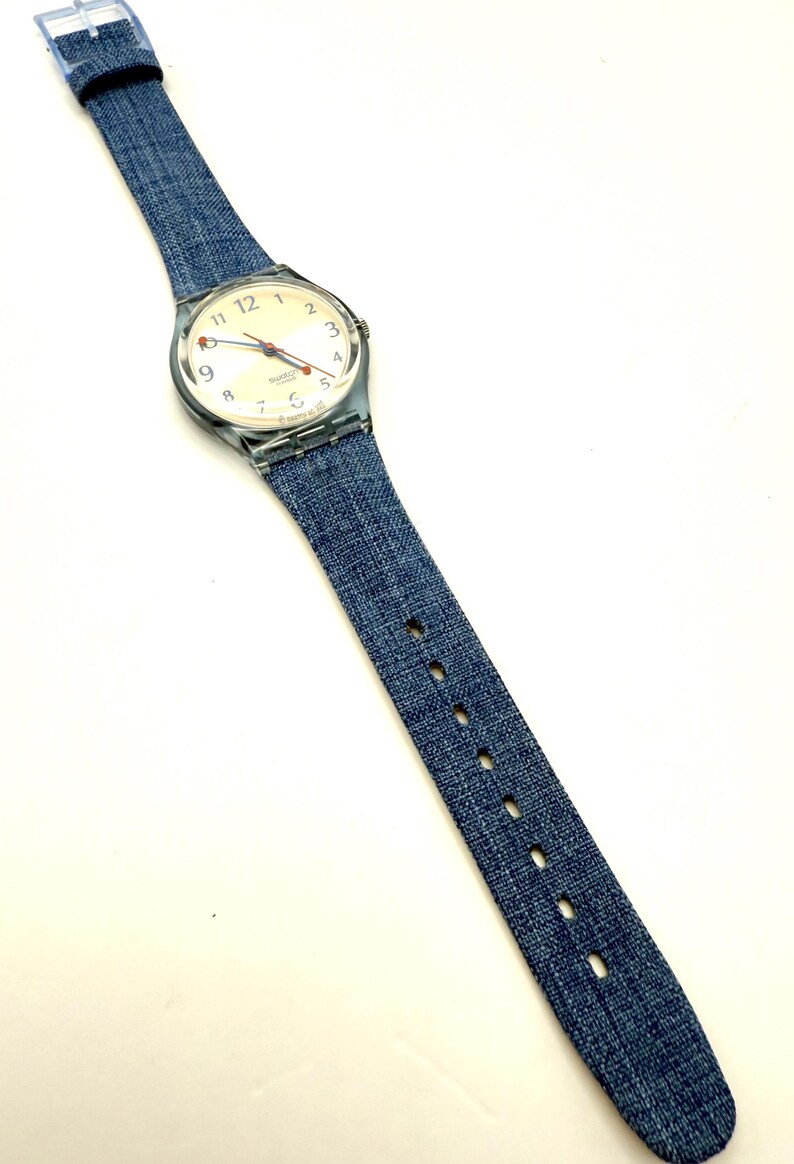 May include: A wristwatch with a clear plastic case and a denim-style blue strap. The watch face is off-white with black numerals and red and blue hands. The strap has a buckle closure and measures approximately 23 cm in length.