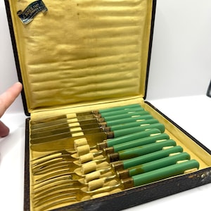May include: An antique set of flatware, including forks and knives, with green handles and gold-colored blades. The set is presented in a vintage, dark brown box with a yellow interior. The box is open, revealing the neatly arranged utensils.
