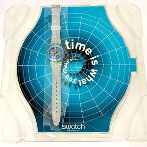 Pu&ograve; includere: Un orologio Swatch con cinturino trasparente e quadrante blu e rosso, esposto nella sua confezione originale. L'orologio &egrave; posto su uno sfondo blu con la scritta "time is what you make of it". Il logo Swatch &egrave; visibile.