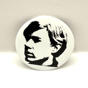 May include: A round, white button featuring a black and white graphic of a person's profile. The image is a stylized portrait with bold lines and shadows, creating a striking contrast against the white background. The button is a simple, yet eye-catching accessory.