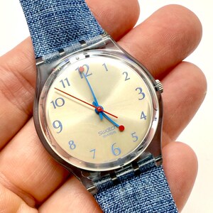 May include: A Swatch Swiss watch with a light-coloured face, blue numerals, and blue and red hands. The watch has a clear plastic case and a blue denim-style strap.